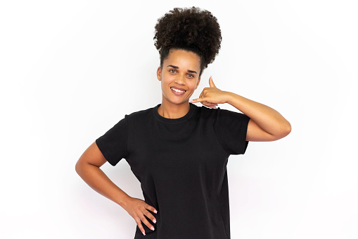 Portrait of happy young woman making call gesture against white background. African American woman wearing black T-shirt smiling at camera and showing phone sign. Contact concept