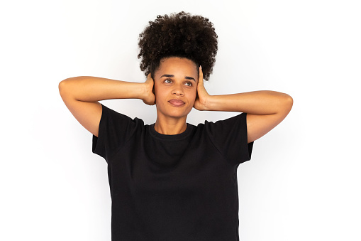 Portrait of ignorant young woman covering ears with hands against white background. African American woman wearing black T-shirt protecting ears from sound. Ignorance concept