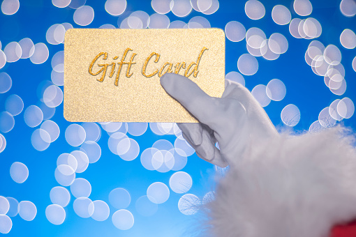 A close-up of Santa Claus’s hand holding a gold glitter gift card against a blue background with white bokeh.