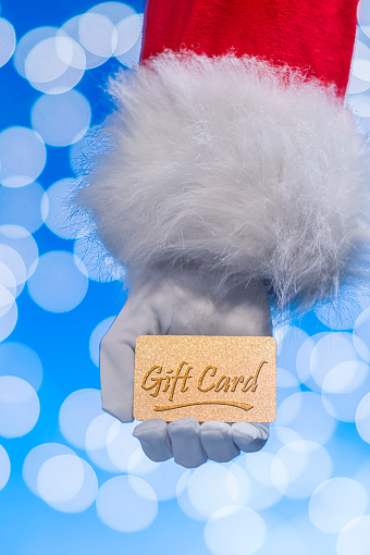 A close-up of Santa Claus’s hand holding a gold glitter gift card against a blue background with white bokeh.