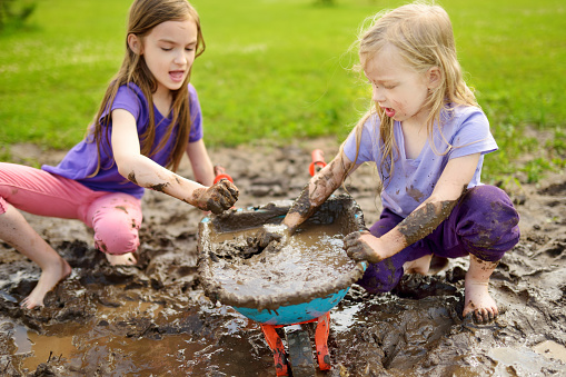 Two funny little girls playing in a large wet mud puddle on sunny summer day. Children getting dirty while digging in muddy soil. Messy games outdoors.