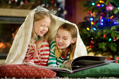 Happy little sisters reading a story book together by a fireplace in a cozy dark living room on Christmas eve. Celebrating Xmas at home.