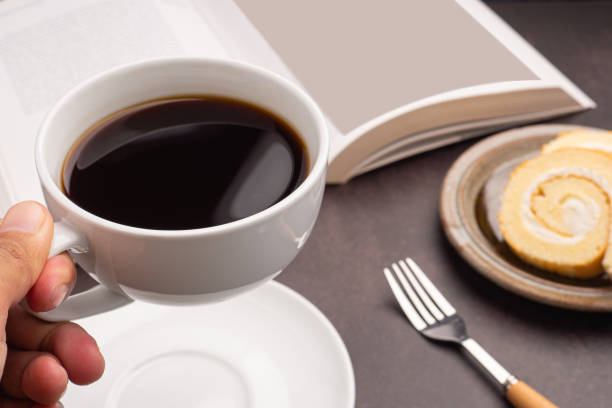 Hand holding a white coffee cup with a book, roll cakes on a plate. Close-up photo. Space for text. Concept of beverage and relaxation.