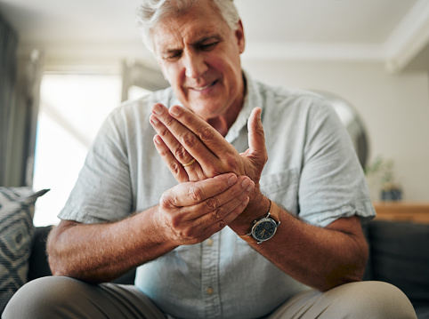 Hands, pain and arthritis with a senior man holding his hand while suffering from osteoporosis, cramp or injury. Health, medical and joint with an elderly male pensioner in the living room at home