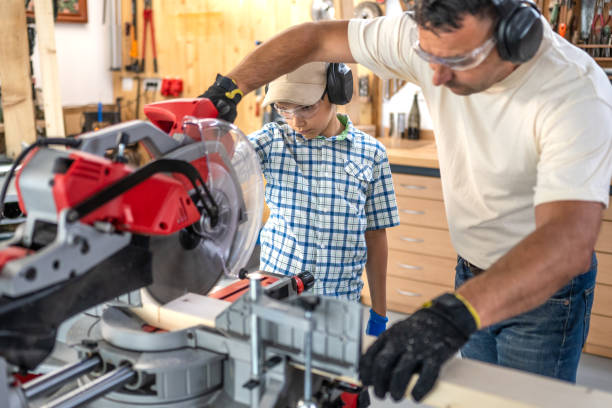 Father and son in protective workwear cutting wood with miter saw, front side view. Craftsman and boy using electrical equipment in carpentry workshop, medium shot