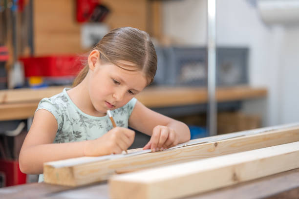 Concentrated girl doing woodwork in craftsman workshop, front side view. Carpentry class for children, kid marking wooden plank with pencil, medium shot