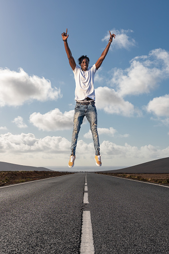Cheerful positive black man enjoying life jumping in the air with raised fists over road in a desert landscape - Travel and enjoying life concept