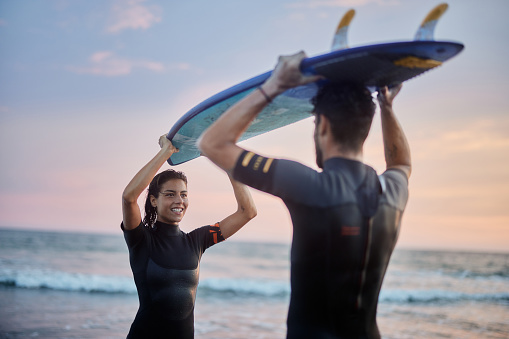 Young happy couple talking while carrying surfboard together on the beach at sunset. Copy space.