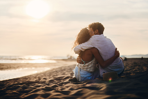 Rear view of happy couple embracing while relaxing in sand on the beach at sunset. Copy space.