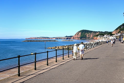 Tourists walking along the promenade with the beach to the left and cliffs to the rear, Sidmouth, Devon, UK, Europe.