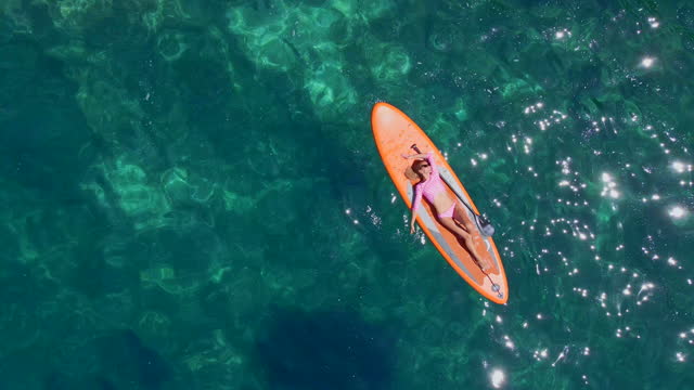 Young woman on a stand up paddle board SUP rawing among beatyful rocks. Clear blue sea water surrounds her. Aerial slowmotion video