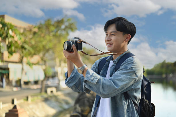 Young hipster man traveler taking pictures with camera while standing on bridge over a river in the city.
