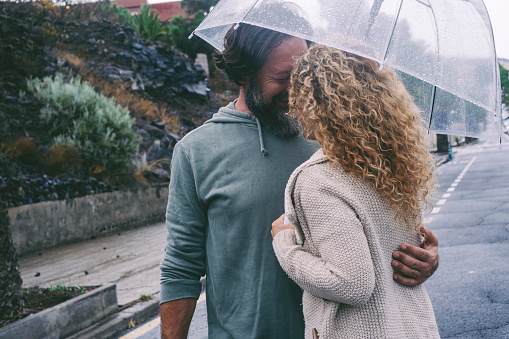 Couple enjoy outdoor leisure activity in rainy day of bad weather hugging and kissing under a transparent umbrella with road street urban in background. Concept of love and relationship man and woman