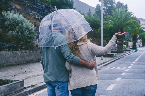 Back view of happy couple in love enjoying rain day with umbrella. Urban street in background. Outdoor leisure activity and concept of relationship together forever. Bad weather winter day. People