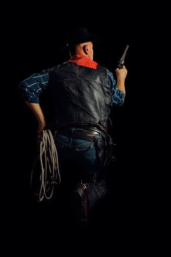 Cowboy is squatting and holding the rope and the gun - back view