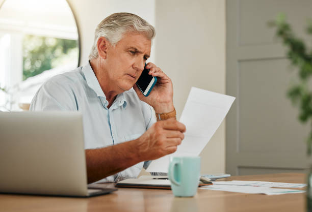 Senior man on his phone with finance documents on call for debt counselling. Old male at home with his laptop and paperwork to check finances, accounting and payments, using his mobile phone for help hombre mayor en una llamada telefónica con documentos financieros para asesoramiento sobre deudas. anciano en casa con su computadora portátil y papeleo para verificar finanzas, contabilidad y pagos, usando su teléfono móvil para obtener ayuda.
