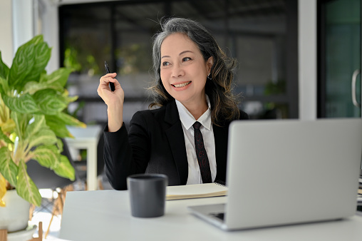 Charming and successful Asian senior female manager or boss sitting at her office desk, smiling, looking out the window, daydreaming about her success.