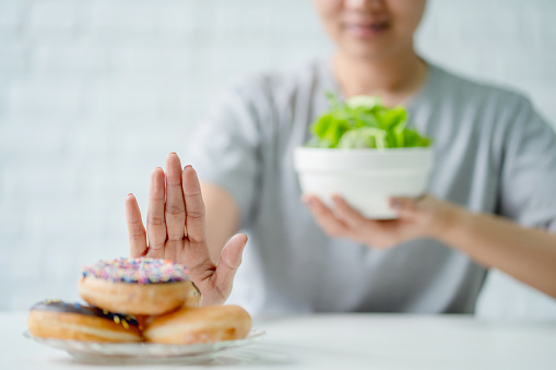 Young Asian woman or girl pushing junk food and sweet donuts with her hands. She chooses salad vegetables for good health.