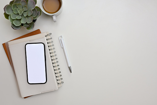Top view, White workspace with smartphone white screen mockup, pencil, spiral notebook, coffee cup, decor plant and copy space.
