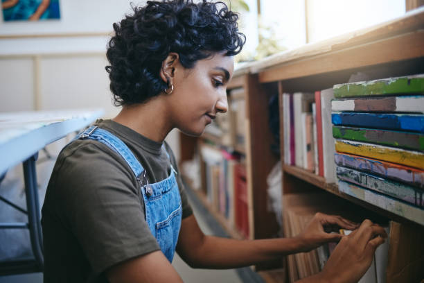 istock Books, creative and Indian student search bookshelf for reading poetry information, learning or knowledge in a work studio or classroom. Teenager woman with history, philosophy or dictionary research 1445392551