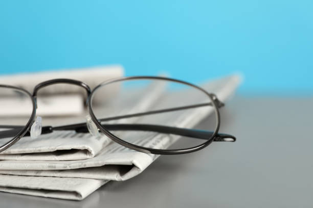 istock Stack of newspapers and glasses on grey table against light blue background, closeup 1445392998