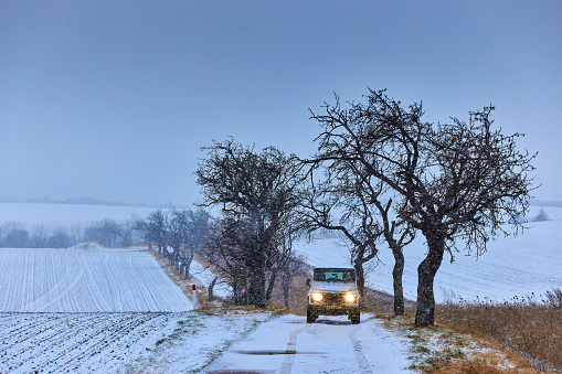 A tree avenue with a lLandrover Defender on Brocken Mountain in Harz region. The lights of the car are on. The sky is grey and the road and the land is covered with snow.
