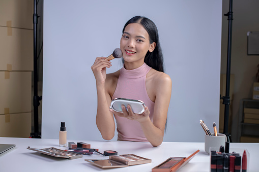 Young Asian woman vlogging herself about beauty products. Woman making a video for her blog on cosmetics looking at camera. Young female blogger holding cosmetics.