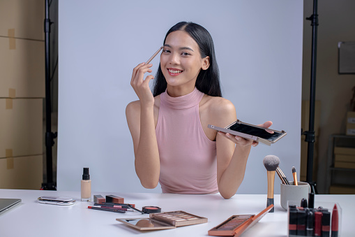 Young Asian woman vlogging herself about beauty products. Woman making a video for her blog on cosmetics looking at camera. Young female blogger holding cosmetics.