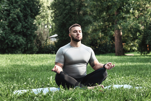 Young bearded athlete man yogi sitting in lotus pose in grass lawn outdoors. Sportsman doing asana yoga in urban park. Male in sportswear meditating, enjoying relaxation, healthy lifestyle. Copy space
