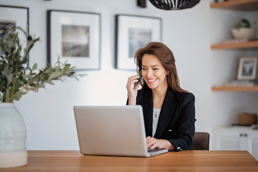 Happy middle aged woman sitting at table and using laptop while working from home