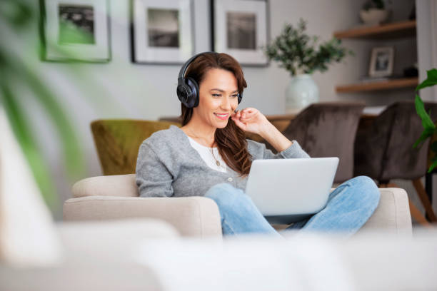 Attractive and cheerful woman using laptop and headphone while sitting in an armchair at home stock photo