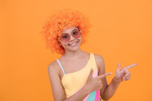 happy child in sunglasses and swimsuit wearing orange curly wig hair pointing finger on orange background, ad.
