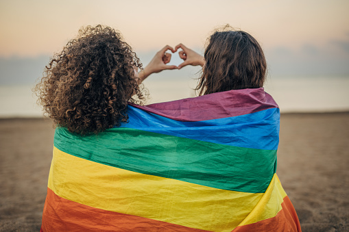 Two women, lesbian couple covered in rainbow flag sitting together outdoors on the beach.