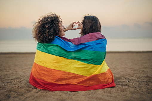 Two women, lesbian couple covered in rainbow flag sitting together outdoors on the beach.