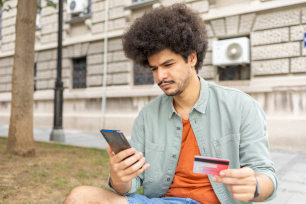 istock Cheerful man sitting on a bench using smart phone and credit card 1445420370