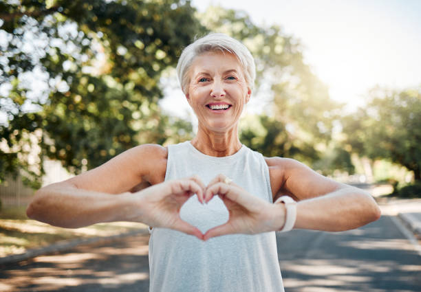 fitness, happy and heart hands of old woman in nature after running for health, wellness and workout. smile, motivation and peace with senior lady and sign for love, faith and training in nature - gezonde levensstijl stockfoto's en -beelden
