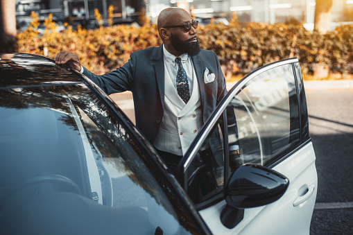 View of a handsome bald black man entrepreneur with a fine black beard, in sunglasses and a tailored fashionable suit, looking aside while entering his contemporary white car parked outdoors