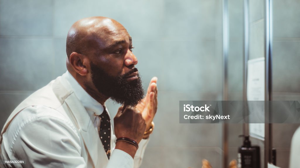 African guy grooming his fine beard A profile view of a handsome bald black man grooming, brushing, and moisturizing the beard hair in front of the mirror in a bathroom of a luxury hotel; an African-American man taking care of his beard Beard Stock Photo