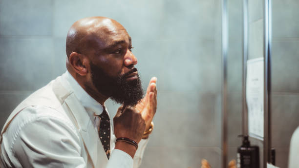 African guy grooming his fine beard A profile view of a handsome bald black man grooming, brushing, and moisturizing the beard hair in front of the mirror in a bathroom of a luxury hotel; an African-American man taking care of his beard beard stock pictures, royalty-free photos & images