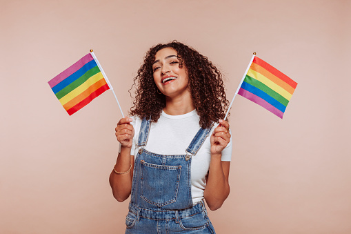 Young cheerful arabian woman wears overalls, holds in hand striped colorful rainbow flags, isolated over beige background. Studio portrait. Homosexual, lesbian, lgtb concept.
