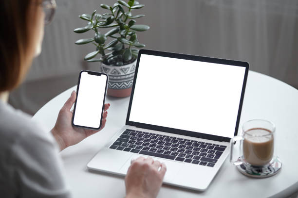 istock Mockup white screen laptop and mobile phone woman using computer while sitting at table at home, back view 1445441455