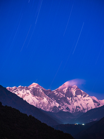 Stars streaking over the alpenglow illuminated peaks of Mt. Everest, Nuptse and Lhotse above the warm lights of Tengboche Buddhist Monastery high in the Himalaya mountains of Nepal.