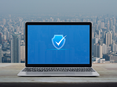 Security shield with check mark icon on modern laptop computer monitor screen on wooden table over city tower and skyscraper, Technology internet cyber security and anti virus online concept