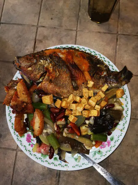 Asian cuisine consists of deep-fried pork and Tilapia (Sea Bass), tofu, underneath sauteed mixed tropical vegetable okra, upo (bottle gourd), talinum or flameflower,  and red beans, served hot on an austere table. Close up