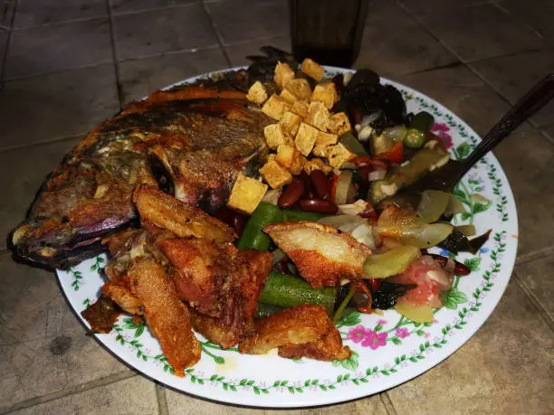 Asian cuisine consists of deep-fried pork and Tilapia (Sea Bass), tofu, underneath sauteed mixed tropical vegetable okra, upo (bottle gourd), talinum or flameflower,  and red beans, served hot on an austere table. Close up