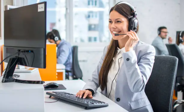 Photo of Friendly female helpline operator in call center. Young woman working in call center and holding microphone on headset with hand.