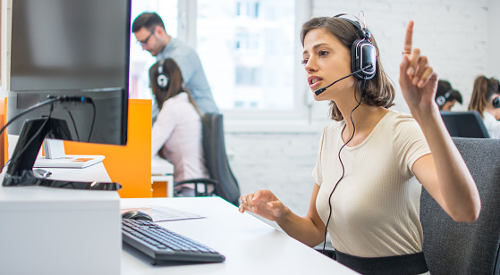 Young female call center operator with hands-free headset talking with client in office.