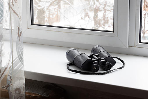 Black binoculars on the white windowsill near a window. The concept of watching and peeping from the window of the house