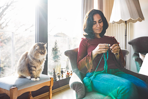 woman sitting in armchair and crocheting, cat beside her on chair is watching