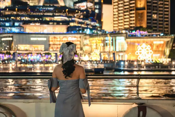 Photo of Beautiful young asian woman in dress looking at illuminated department store glowing in festival event on riverside in the night. Dinner cruise sailing and sightseeing on the Chao Phraya river